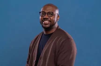Corey Norman smiling, wearing glasses and a brown cardigan against a blue background