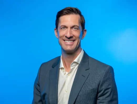 Brian Boosi smiling, wearing a dark gray suit jacket and a white open-collar shirt against a bright blue background